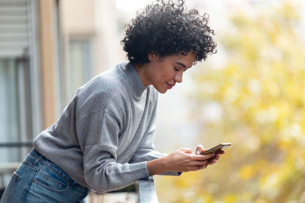 A cheerful young black girl is talking on the phone to a friend while wearing a gray sweater and blue jeans.