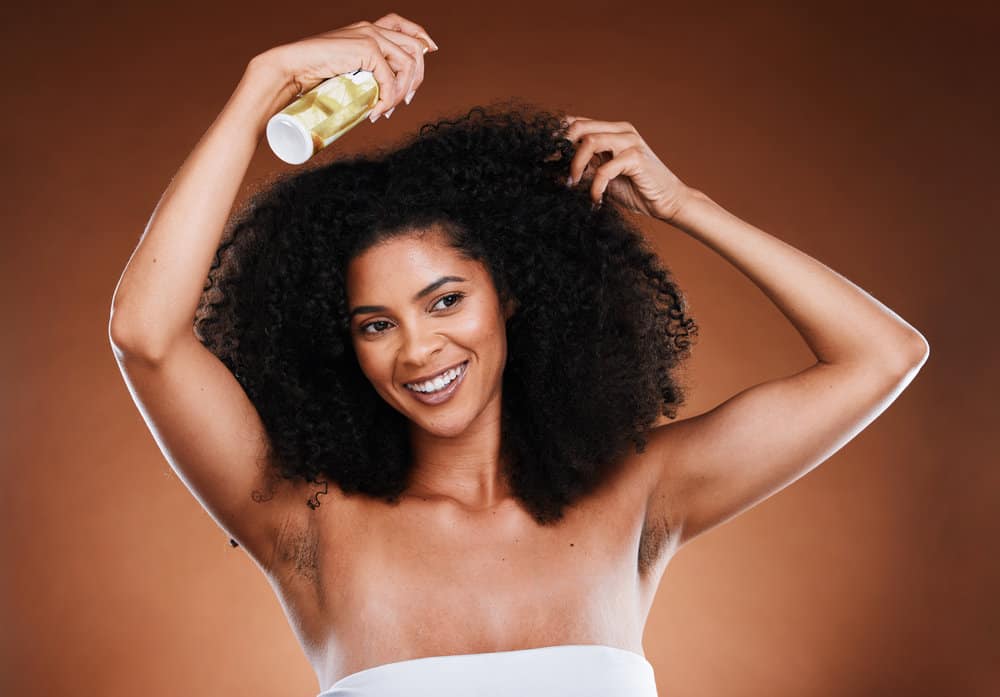 A black female is spraying her hair shaft with a leave-in conditioner - a stable in her hair care routine.