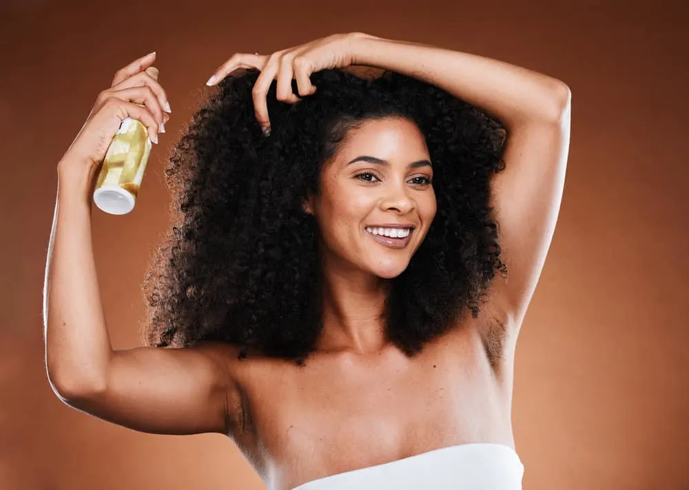 A female with curly hair that's concerned about hair loss is spraying her dry hair with a mask-like creamy product.