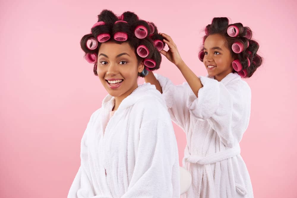 A parent and kid with long hair and different curl patterns using different-sized rollers to prep hair with tight ringlets.