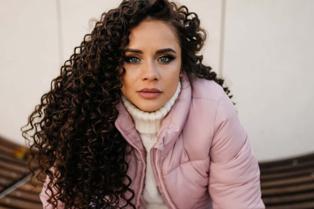 A confident female with a long hair perm is sitting outside wearing a cute purple jacket and white sweater.