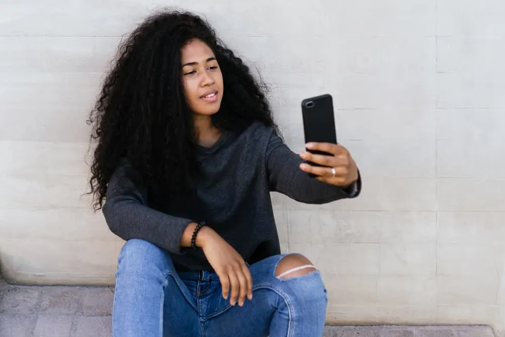 A young American female with curly locks is admiring her healthy hair while taking a selfie photo.