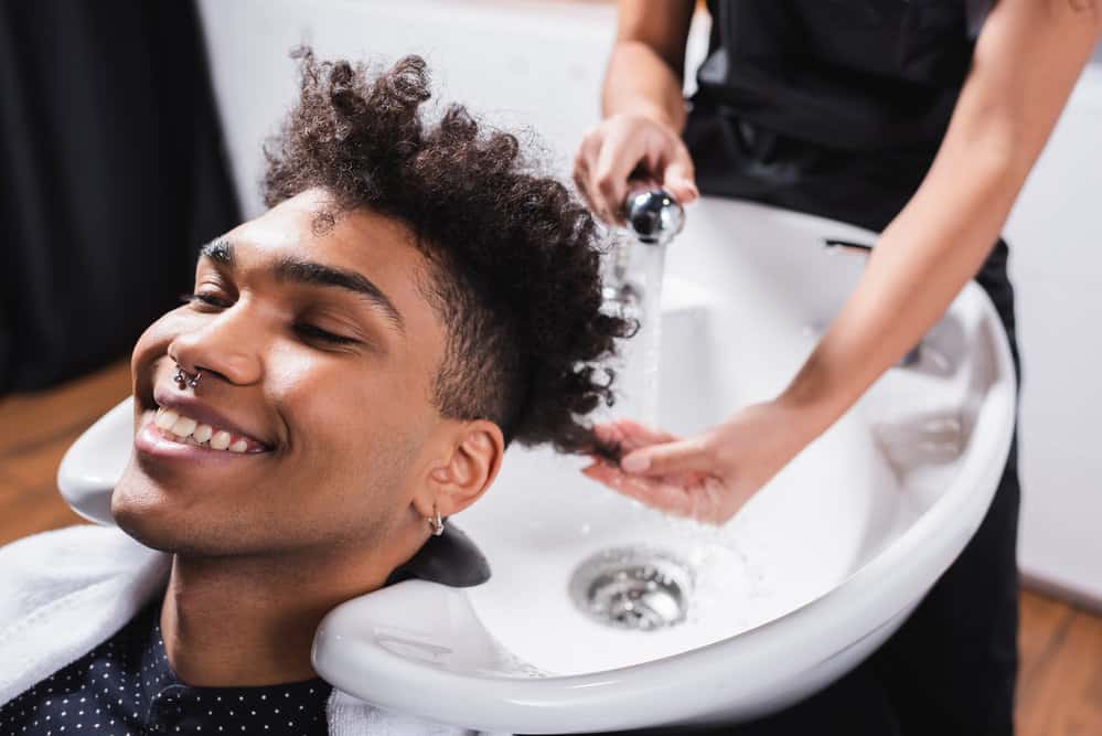 A black guy with naturally curly hair is getting a shampoo and conditioning treatment before a haircut appointment.