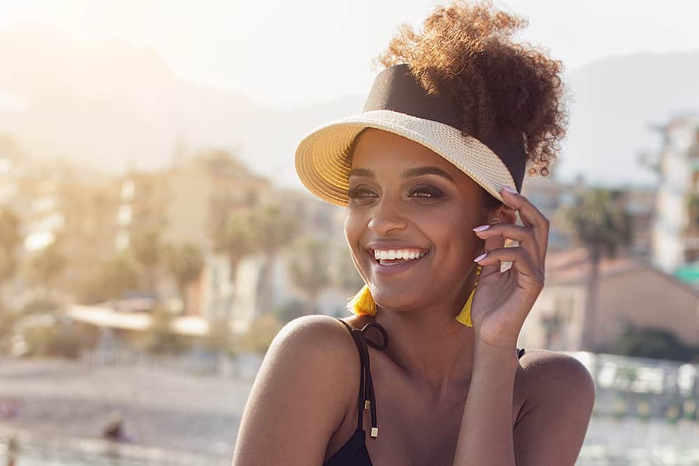 A stylish African American female with swimmers hair sits on the beach wearing a black swimsuit and yellow earrings.