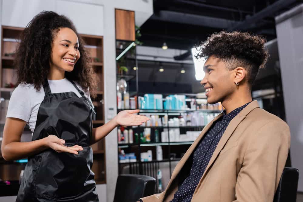 A man is sitting in a salon chair discussing his next hair appointment and about to give his professional hairstylist a tip.