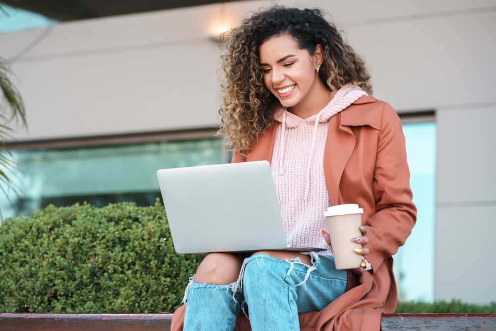 A Latin female with fine hair uses her laptop to determine how to repair hair shaft damage on her unwashed hair.