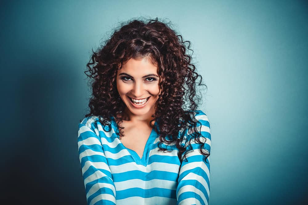 A cute white female with a great smile has thick hair with tighter curls created with a DIY sock bun and hair ties.