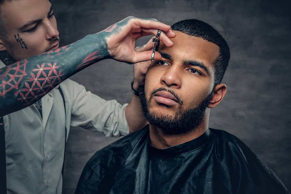 A stylish tattooed barber creates a great haircut on a black man with longer hair that takes roughly 45 minutes.