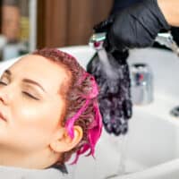 A cute white lady is getting pink and red hair dye done in a porcelain sink by a professional wearing rubber gloves.