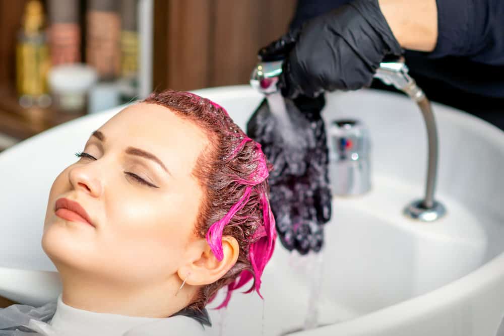 A cute white lady is getting pink and red hair dye done in a porcelain sink by a professional wearing rubber gloves.