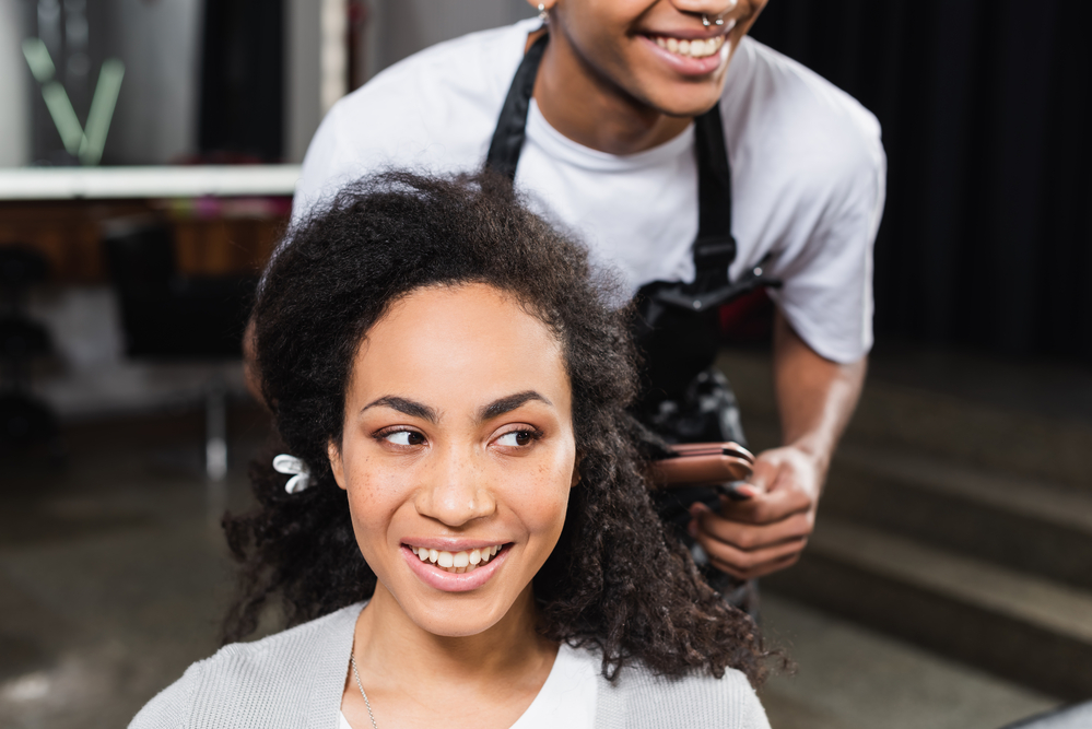 A black woman with naturally curly hair is getting a flat iron hairstyle at a local hair salon with many hairdressers.