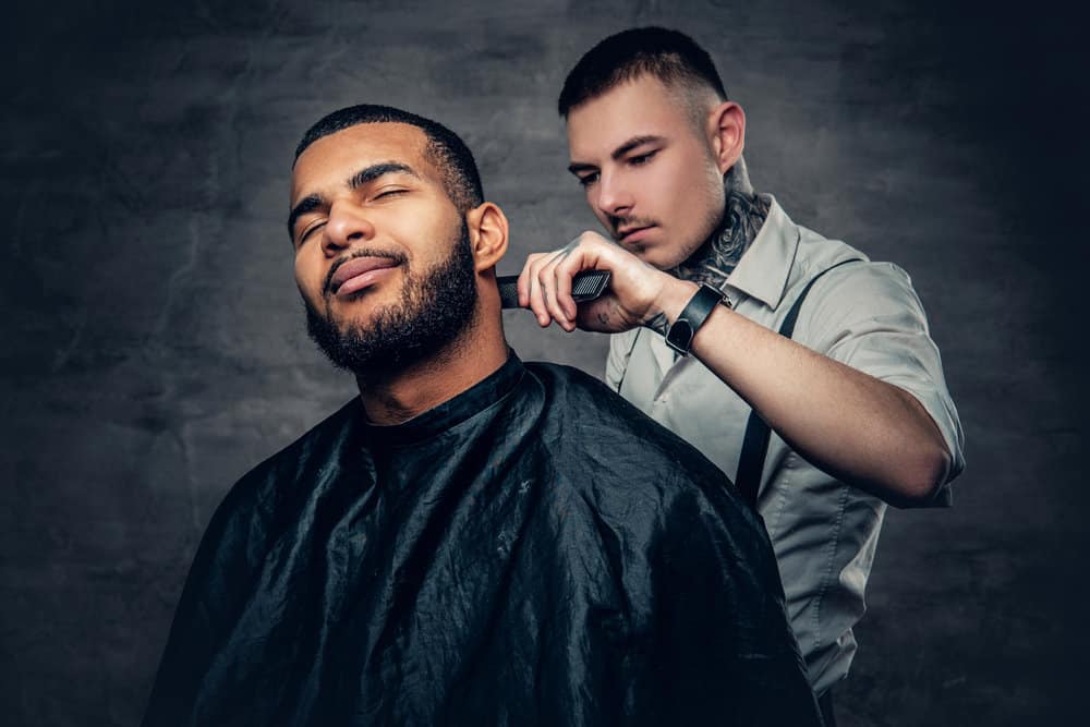 A light-skinned black dude wearing a dark-colored haircutting cape while getting a professional shave. 
