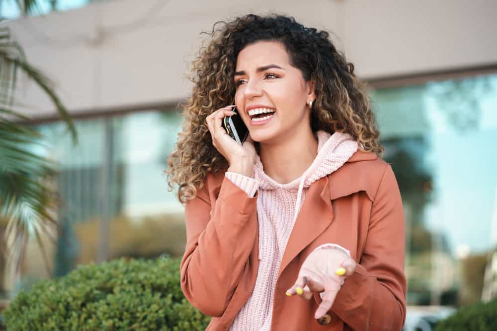 A joyful female with long hair and beautiful yellow fingernails is discussing her oily hair with a friend over the phone.