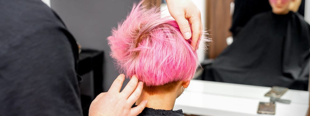 A hairstylist is finishing up a hair dyeing session from a client wearing a new hot pink color on a stylish bob hairdo.