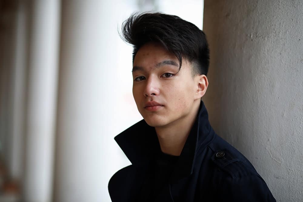 A young adult male is wearing popular Korean messy hair that has been brushed forward with a wavy texture.