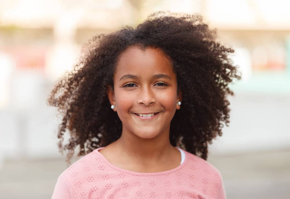 A cute African American girl wearing a simple hairdo with the cutest white pearl earrings and a pink sweater.