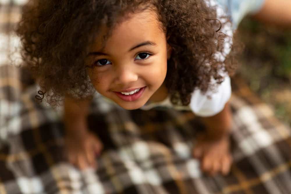 A cute little girl playing indoors as her mom prepares to braid curvy cornrows with white beads for a special event.