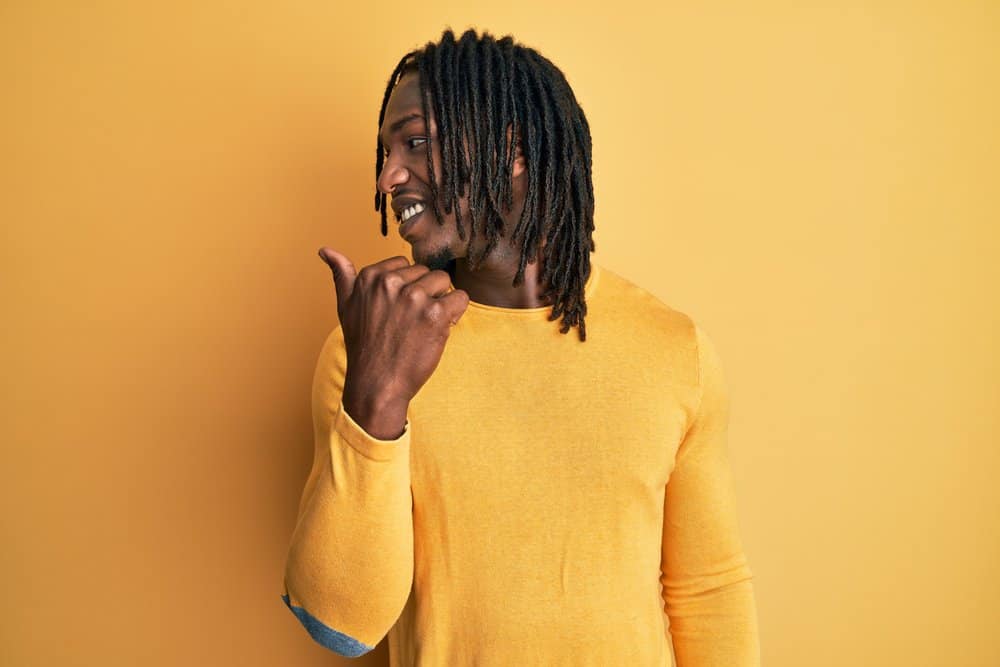 A black man with long freeform dreads is wearing a yellow form-fitting muscle shirt while looking behind him.