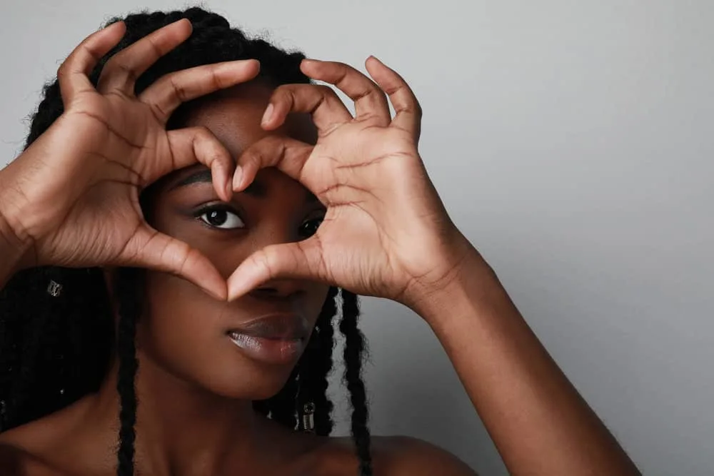 A black girl with dark brown hair strands making a heart symbol with her hands covering her right eye.