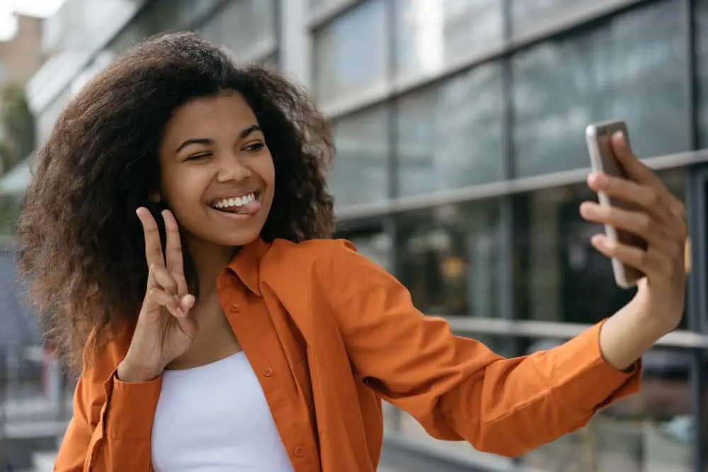 A black girl wearing a pure silk shirt while making a peace sign with her right hand and taking a selfie for a friend.