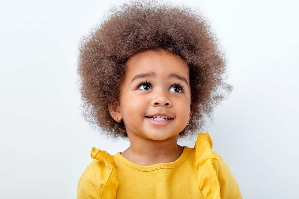 A precious baby girl with a curly, full afro hairstyle and a sweet expression highlighting beautiful gold earrings.