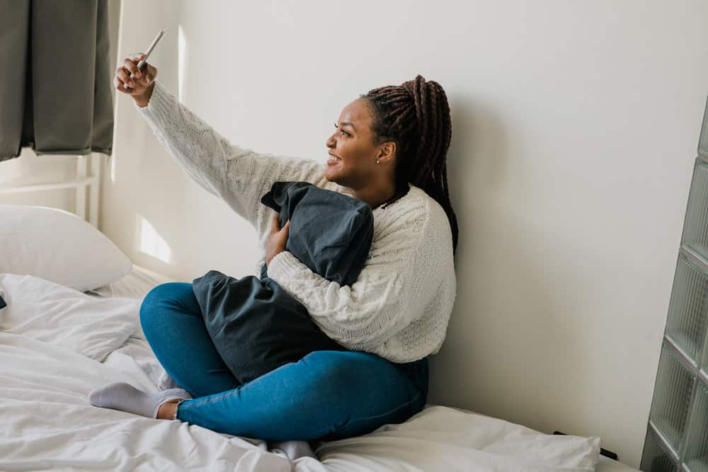 African American female with box braids taking a selfie with her iPhone while sitting on her bed.