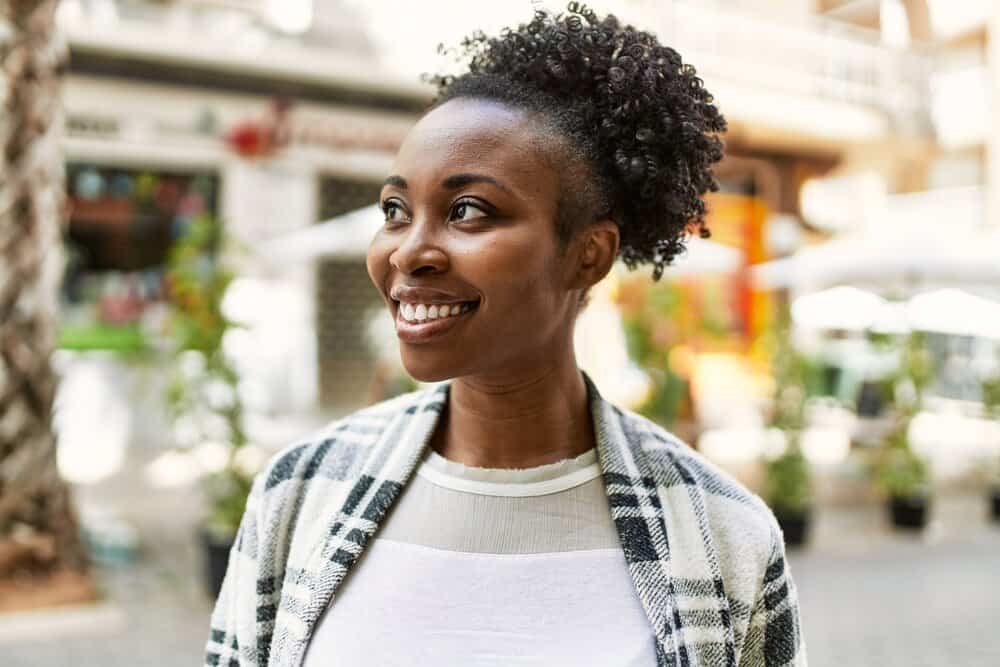 A black woman with dark brown henna-dyed hair is wearing an old t-shirt and a black and white plaid jacket.