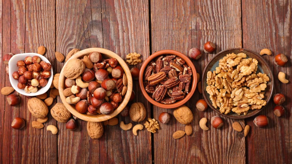 A colorful and textured assortment of various nuts, including almonds, walnuts, pecans, and cashews, displayed together in a rustic wooden bowl.