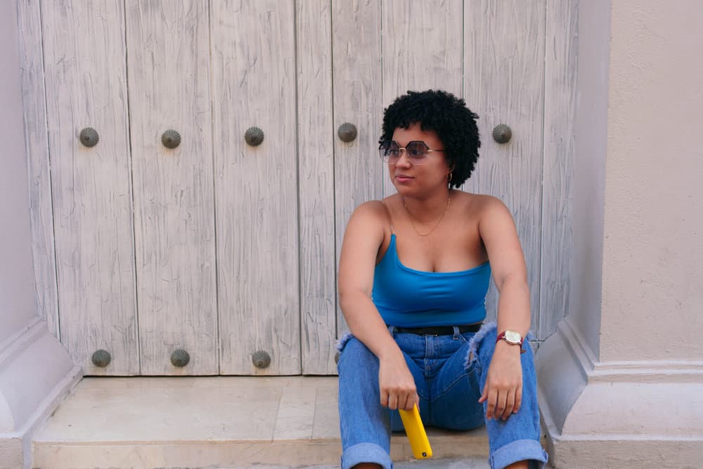 A charming black woman with thing fine hair sits on a step outside while holding her cell phone.