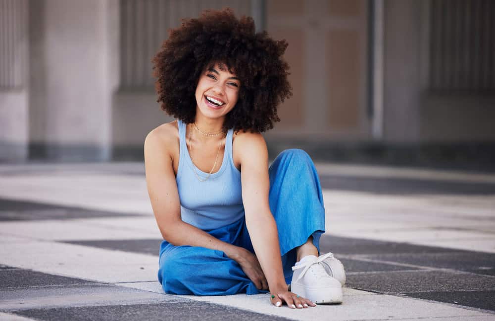 A cute black female is sitting on the floor with a trendy style that uses hydrolyzed wheat protein on her natural curls.