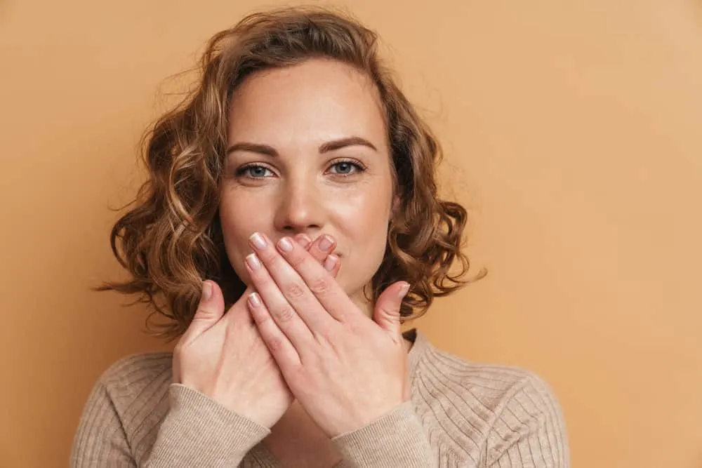 12 Best Medium-Length Haircuts With Layers, Bangs, & More 19 A cute white girl with mid-length hair and fine hair strands is covering her mouth with her hands like she's surprised.