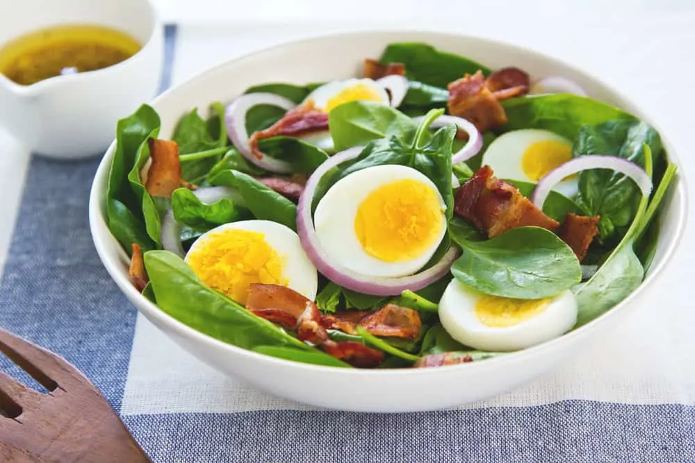 A delicious and hearty salad consisting of crispy bacon, a boiled egg, and a bed of fresh spinach leaves topped with a tangy salad dressing.