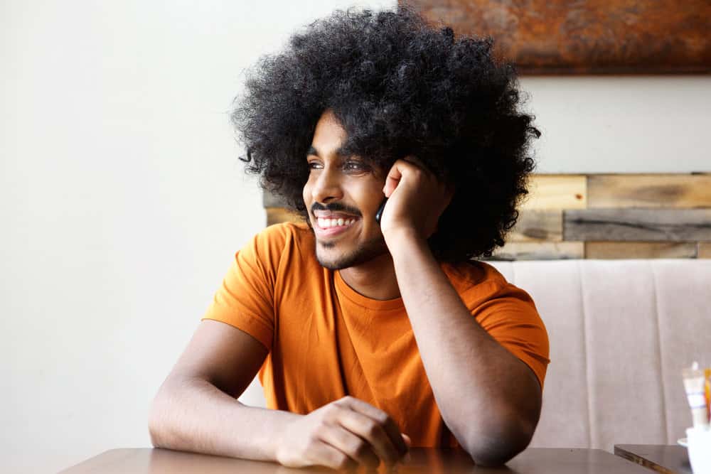 A cheerful African American man with long hair styled with castor oil and tea tree oil that's sitting in a coffee shop.