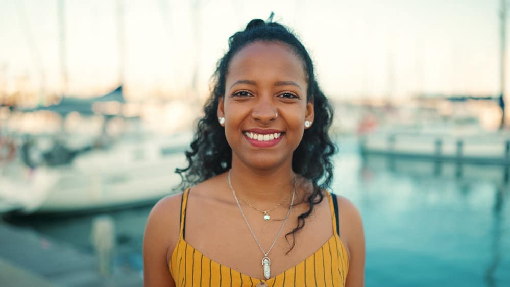 A cute black woman with a dazzling grin has long curly natural hair while wearing custom makeup and lipstick.