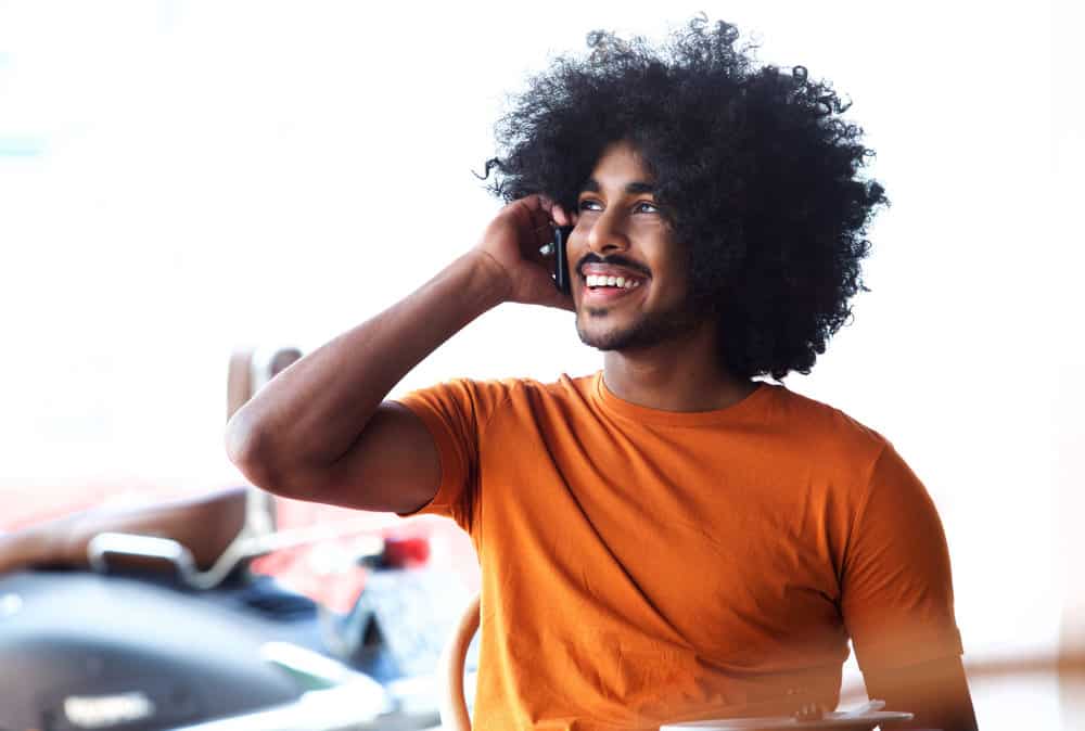 A handsome young black man with thick hair talking to a friend on the phone about hair growth tips and scalp health.