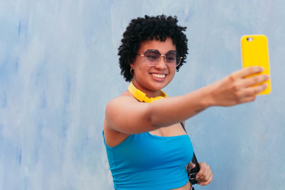 A cute black girl with naturally curly hair is taking a selfie of her type 4 hair texture with face-framing layers.