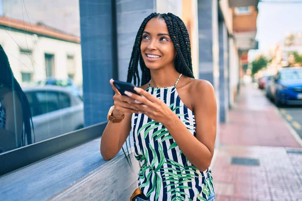 Black woman with Ghana braids wearing a leather watch, flower-based shirt, and blue jeans while using her iPhone 10.