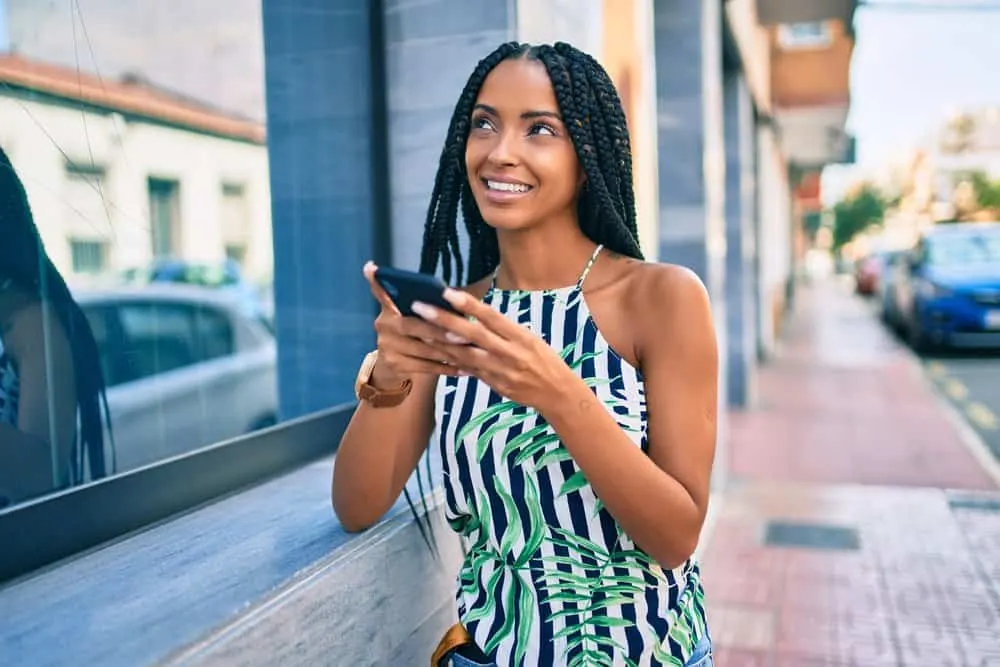 Black woman with Ghana braids wearing a leather watch, flower-based shirt, and blue jeans while using her iPhone 10.