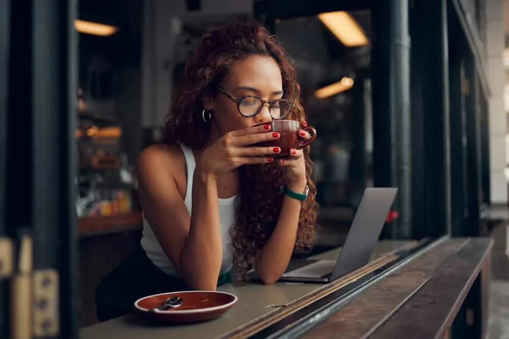 Why Is My Hair Flat and Greasy? How to Fix Flat Greasy Hair 8 An African American female sitting in a coffee shop researching the scalp's oil production on her laptop.
