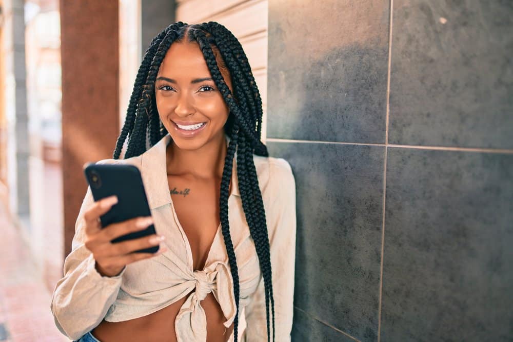 Female wearing box braids leaning against a tile while, talking on a mobile phone.