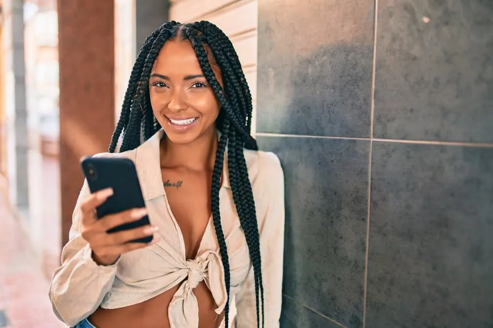 Female wearing box braids leaning against a tile while, talking on a mobile phone. 
