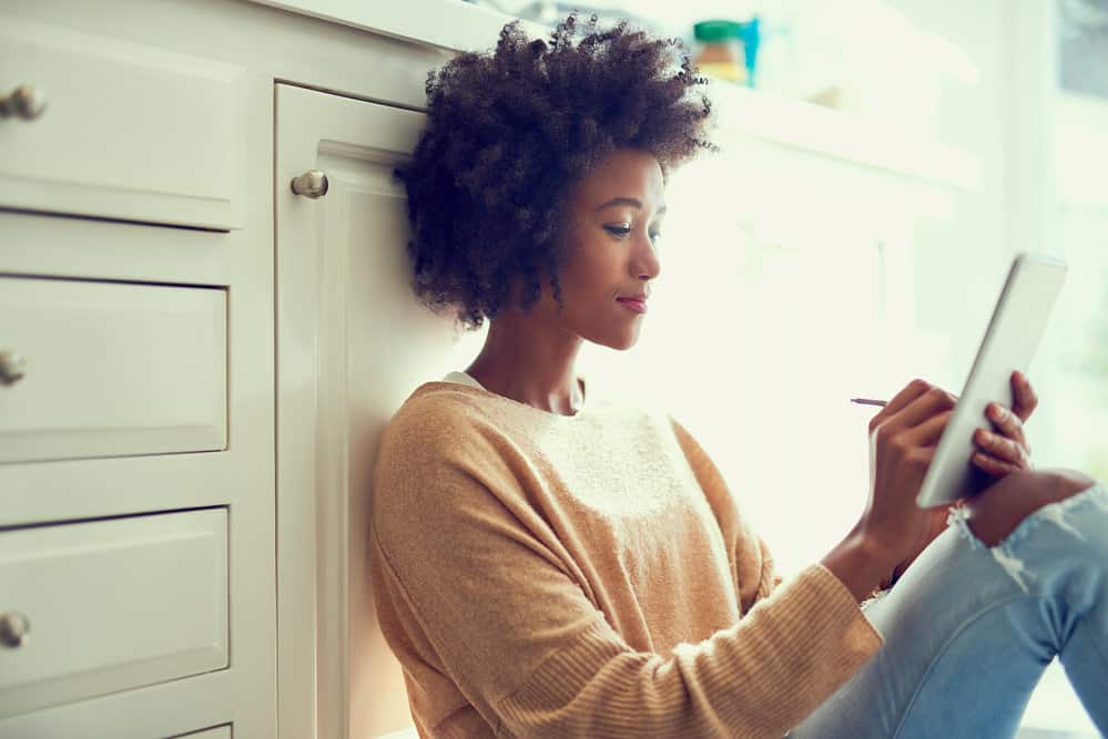 A young female sitting on the kitchen floor using an iPad to read an article about hair frizz.