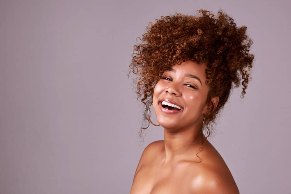 A gorgeous black female with long curly hair showcases a natural curl pattern, styled in a messy bun with a few curls left loose for an effortless style.