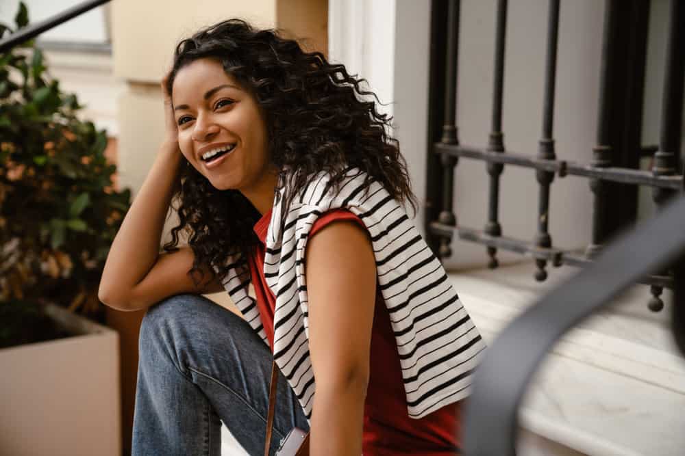 The fashionable young black woman embraces her 3B natural curl pattern, smoothing frizzy curls with nourishing hair oil to mitigate breakage.