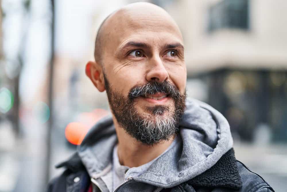 A young man with thinning hair follicles is sporting a bald head with grey hair in his thick facial hair.