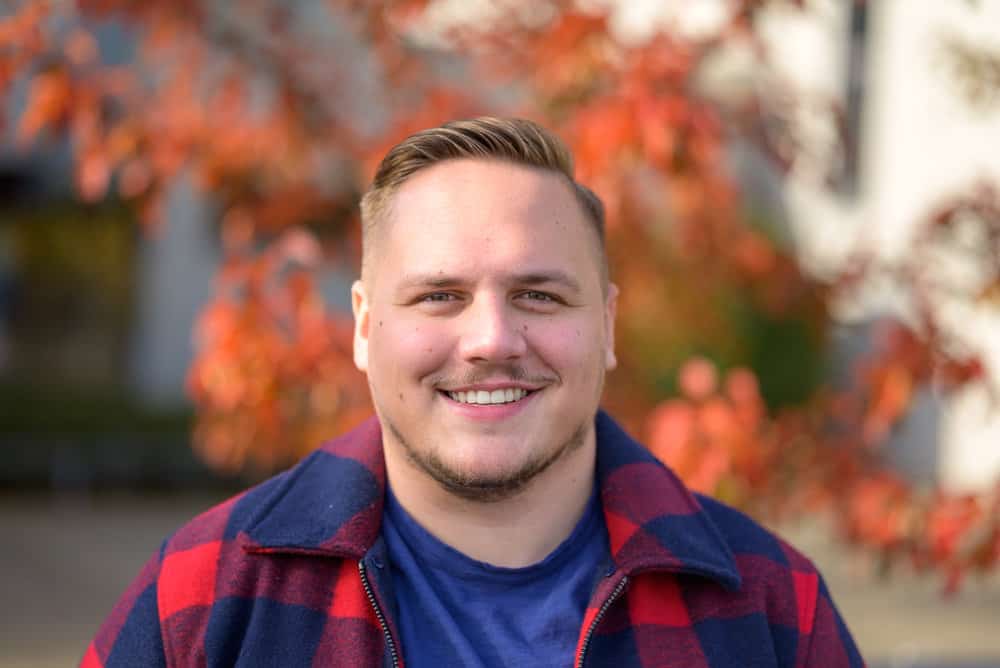 A young man with a round face shape has thinning facial hair and a high and tight fade, which is one of our favorite hairstyles for round faces.