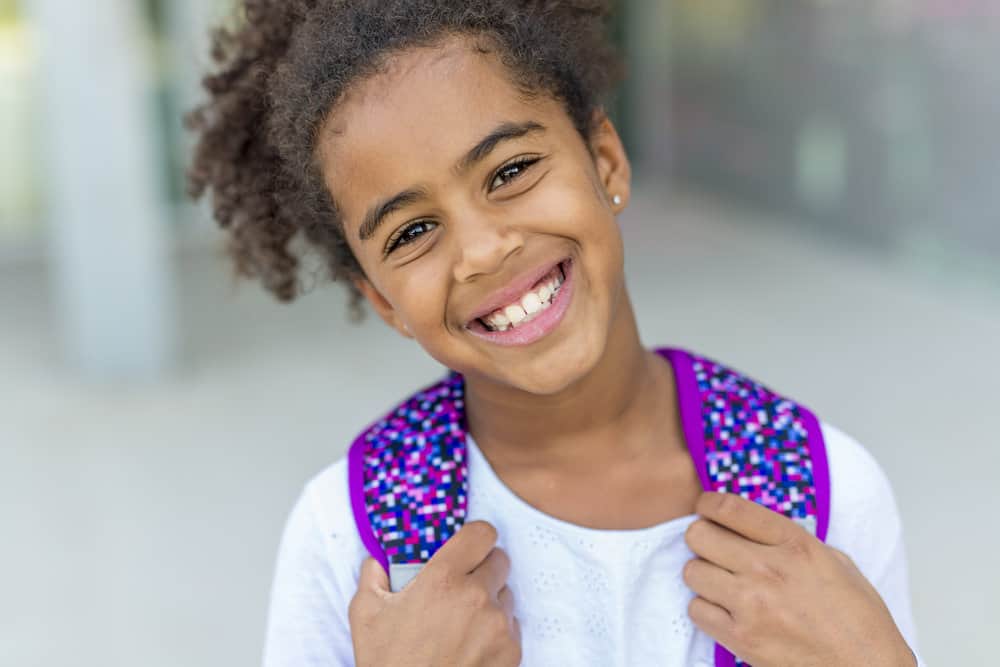 A cute young black girl with dark brown, thick hair in a simple bun with curly ends is wearing a classic style that her mom created with human hair extensions.