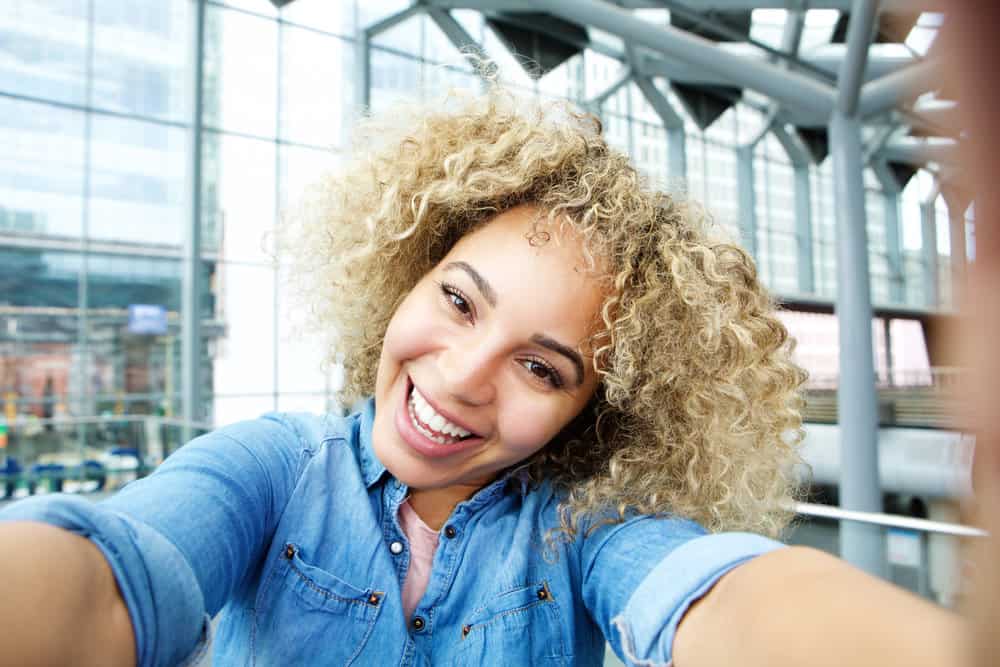 A joyful young African American female with blonde hair and light brown undertones following a hair bleach treatment by a professional colorist without any yellow or brassy tones.