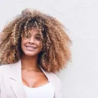 A stunning black female with a type 3 curl pattern styled with natural oils has beautiful blonde and brown hair strands creating a cool balayage look on her natural curls.