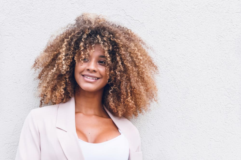 A stunning black female with a type 3 curl pattern styled with natural oils has beautiful blonde and brown hair strands creating a cool balayage look on her natural curls.